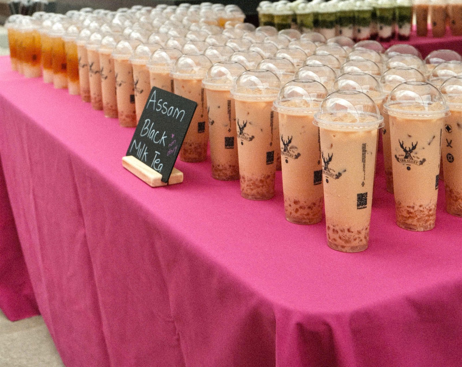 Rows of Assam Black Milk Tea cups on a pink-draped table with a chalkboard sign